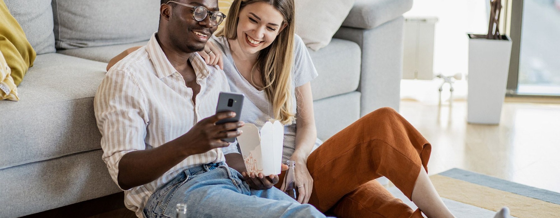 a man and woman with take out, sit against a couch on their living room floor and watch at their cell phone